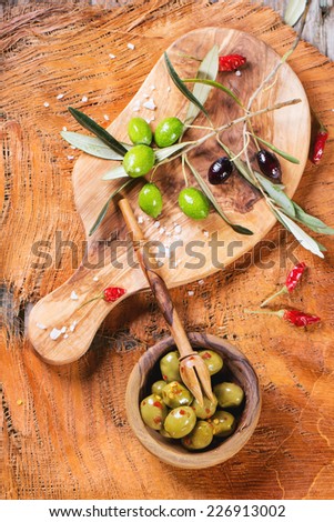 Green olives in olive wood bowl with chili pepper and olive\'s branch over wooden table. Top view.