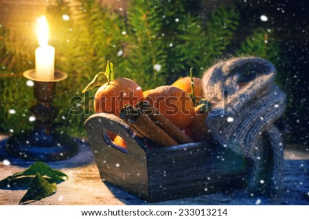 Wooden basket with tangerines, cinnamon sticks and scarf over wooden background with burning candle, snow and Christmas tree.