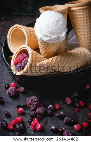 Homemade vanilla ice cream in wafer cones and empty waffer cones, served in metal bowl with frozen berries and spoon over black table. See series