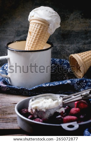 Vanilla ice cream in wafer cones and empty wafer cones, served in metal bowl with frozen berries and spoon over wooden table with blue textile