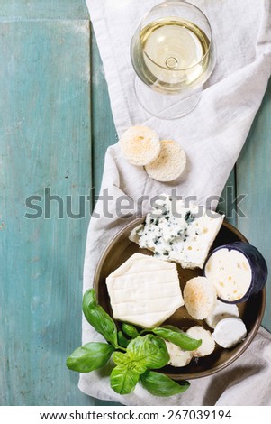 Variation of cheese, served with fresh basil, bread and glass of white wine over turquoise wooden table. Top view. See series