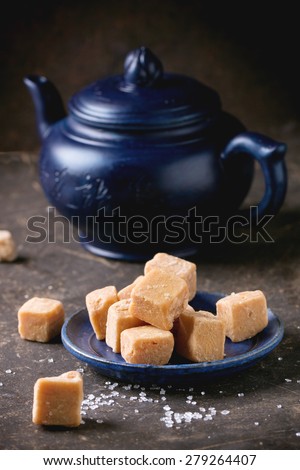 Blue ceramic plate with fudge candy ceramic teapot, served with sugar cubes over dark background.