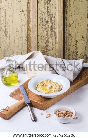 Plate of homemade hummus with bread, olive oil and raw chick-pea, served on wooden cutting board over rustic table with gray tablecloth