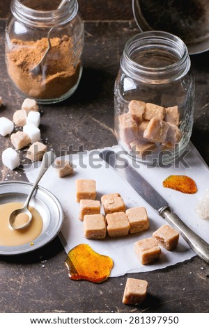 Fudge candy and caramel on baking paper and in glass jar, served over dark background with plate of caramel sauce and sugar cubes. See series