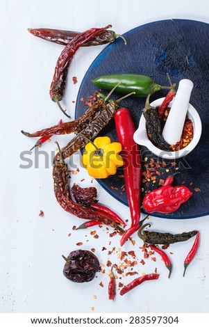 Assortment of fresh, dryed and flakes hot chili peppers with white ceramic mortar on dark blue cutting board over light blue wooden background. Top view