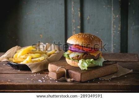 Fresh homemade burger on little cutting board with grilled potatoes, served with ketchup sauce and sea salt over wooden table with gray wooden background. Dark rustic style.