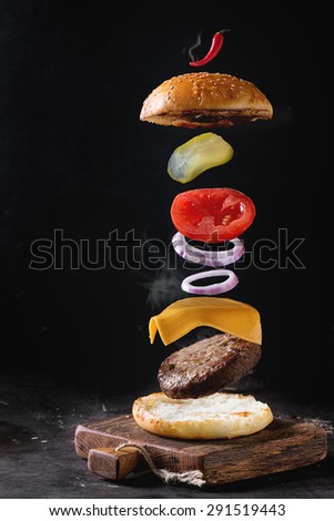 Flying ingredients for homemade burger on little wooden cutting board over dark background.