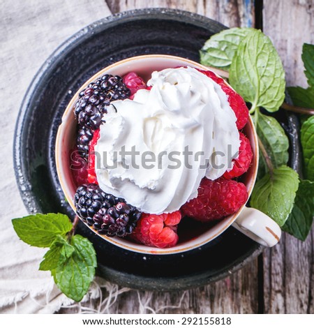 Cup of raspberries and blackberries served with fresh mint and whipped cream over old wooden table. Top view. Square image