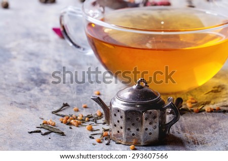 Vintage tea strainer as teapot and glass cup of hot tea with dry green tea leaves. Old tin background.