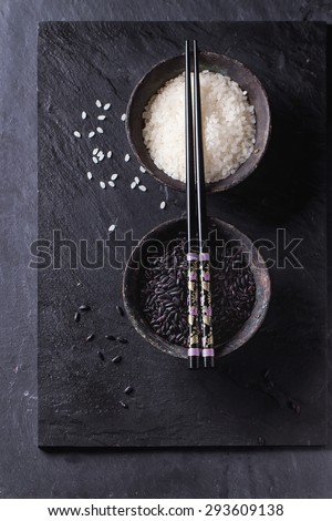 Black and white rice in old metal china bowls with black chopsticks over black slate background. Top view.