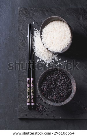 Black and white rice in old metal china bowls with black chopsticks over black slate background. Top view