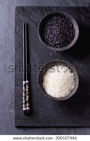 Black and white rice in old metal china bowls with black chopsticks over black slate background. Top view