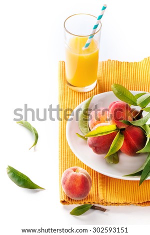 Peaches on branch with leaves in white ceramic plate and glass with peach juice with cocktail tube over orange textile napkin. Over white background.
