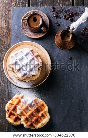 White plate with homemade belgian waffles with sugar powder, ceramic cup of coffee, old cezve and coffee beans. Over black slate as background. With copy space at right. Top view.