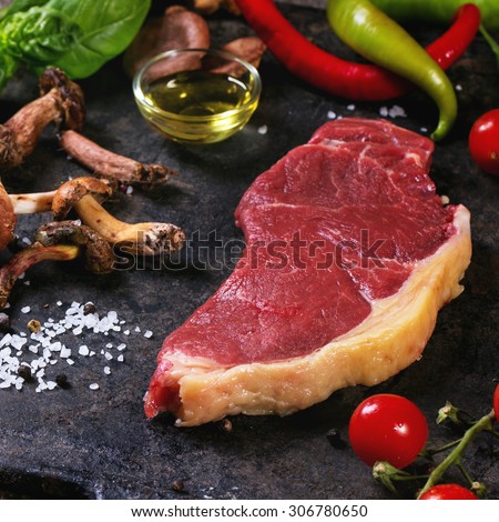 Raw steak served with vegetables and forest mushrooms on black metal cutting board over old wooden table. Square image with selective focus