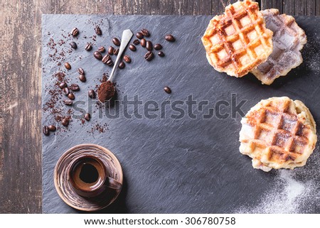 White plate with homemade belgian waffles with sugar powder, ceramic cup of coffee, teaspoon and coffee beans. Over black slate as background. With copy space at centre. Top view.