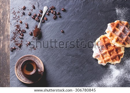 White plate with homemade belgian waffles with sugar powder, ceramic cup of coffee, teaspoon and coffee beans. Over black slate as background. With copy space at centre. Top view.