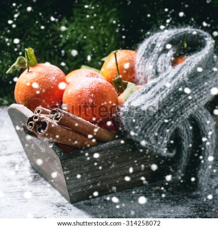 Wooden basket with tangerines, cinnamon sticks and scarf over wooden background with snow and cone. Square image with selective focus