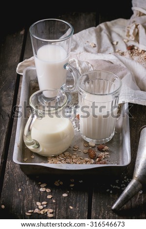 Set of non-dairy milk (rice milk, almond milk and oat milk) in glass cups and jug on old aluminum tray with rice grains, oat flakes and almond over old wooden table. Dark rustic style.
