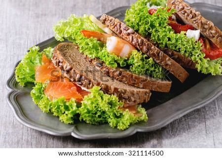 Set of Sandwiches with whole wheat bread, fresh salad, feta cheese, cherry tomatoes, shrimp and salted salmon on vintage metal plate over white wooden table.