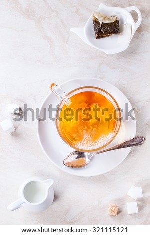 Glass cup of hot tea on saucer with sugar cubes, jug of milk and tea bag over white marble backgtound. Top view