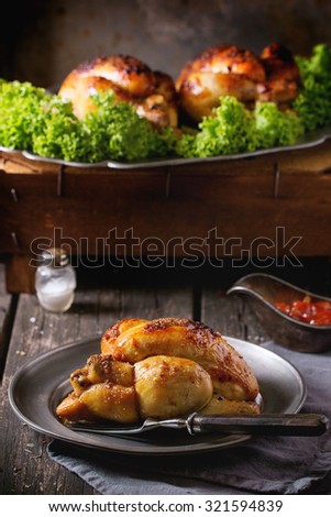 Whole Grilled mini chicken on vintage metal plate and dish with chicken and green salad behind over old wooden table. With fork, napkin and spices. Dark rustic style.