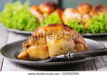 Whole Grilled mini chicken on vintage metal plate and dish with chicken and green salad behind over whitw wooden table.