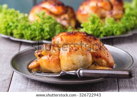 Whole Grilled mini chicken on vintage metal plate and dish with chicken and green salad behind over whitw wooden table.