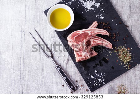 Raw lamb chops with salt, pepper, dry herbs and bowl of olive oil on black slate board over white wooden table. Meat fork near. Top view. With copy space at left