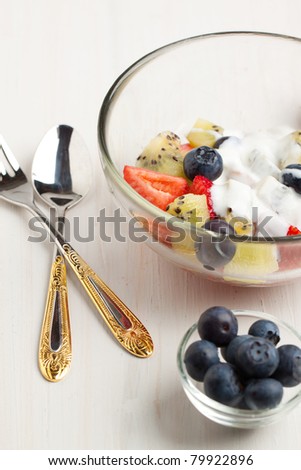 Glass bowl of fruit salad with fresh strawberries, blueberries and kiwi with whipped cream, silver dinner set on white table
