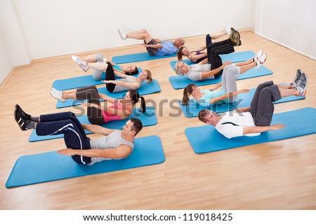 Large group of diverse people exercising in a gym class lying on blue mats doing leg raising and twisting exercises