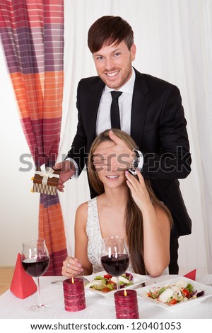 Handsome young man surprising his wife or sweetheart with a gift as she sits at a stylish restaurant table enjoying a romantic meal