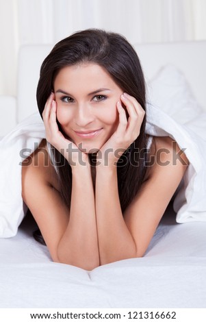Smiling woman lying on her stomach on her bed under the bedclothes resting her chin on her hands