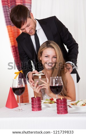 Handsome young man surprising his wife or sweetheart with a gift as she sits at a stylish restaurant table enjoying a romantic meal