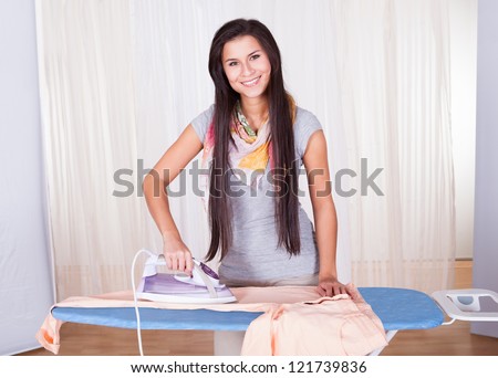 Cheerful housewife with a beautiful smile standing at the ironing board ironing clothes against a white curtained window with copyspace
