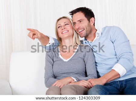 Laughing couple sitting close together on a couch pointing off screen to the left and looking in that direction