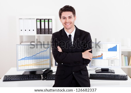 Confident smiling stock broker sitting on the edge of his desk surrounded by graphs and analytics indicating a successful bull market