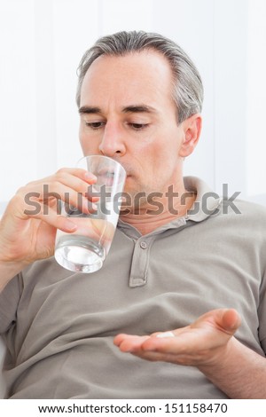 Ill man taking medication sitting up holding a glass of water looking at a tablet in her hand