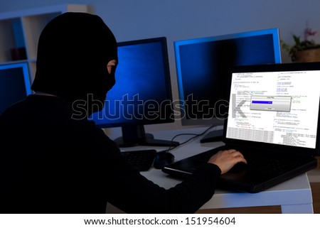 Masked hacker wearing a balaclava sitting at a desk downloading private information off a computer