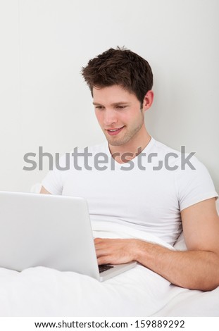 Young Man Using Laptop Sitting On Bed In Bedroom