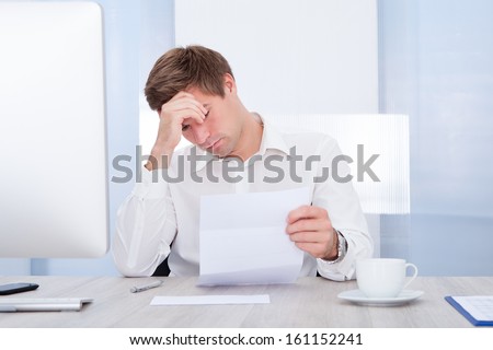 Frustrated Businessman Sitting At Office Desk Holding Paper