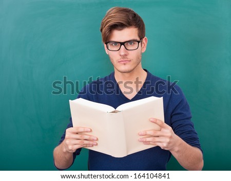 Portrait Of A Young Student Holding Book In Front Of Green Chalkboard