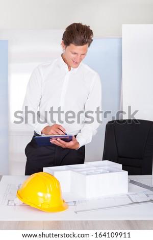 Male Architect Looking At Architectural Model Writing On Clipboard