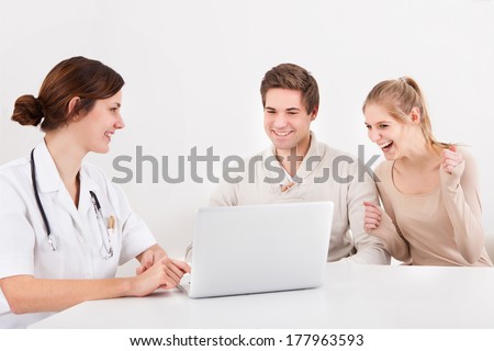 Excited Couple Looking At Laptop With Doctor Sitting At Desk