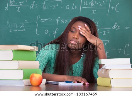 Tired female teacher with hand on head sitting at classroom desk