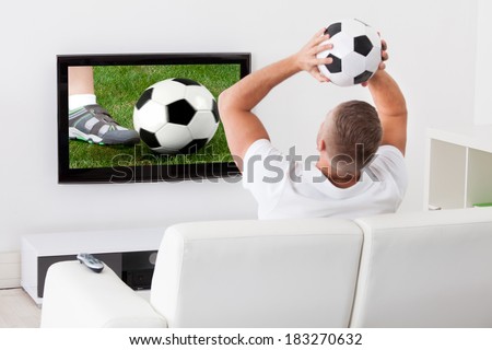 Excited soccer fan watching a game on television holding a soccer ball above his head as he sits on a comfortable sofa in his living room