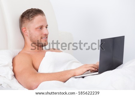 Man sitting up in bed against the pillows working on a laptop computer smiling as he reads the screen