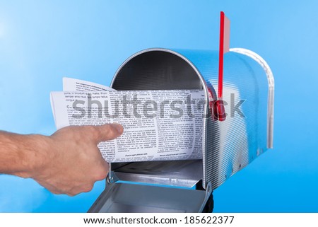 Man removing a newspaper from his mailbox with a closeup of his hand clasping the folded paper against a blue sky