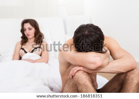 Sad shirtless man sitting on bed with woman in background at home