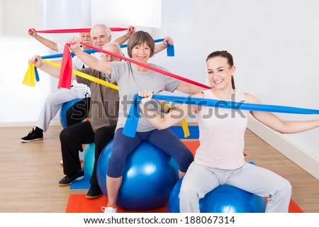 Portrait of trainer and senior customers with resistance bands sitting on fitness balls at gym